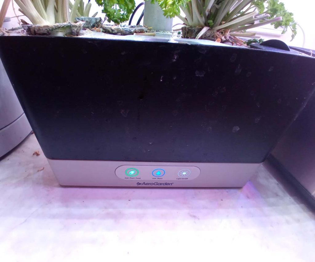 Front view of a black AeroGarden Harvest water tank base with illuminated control buttons and visible mineral residue on the outer shell.