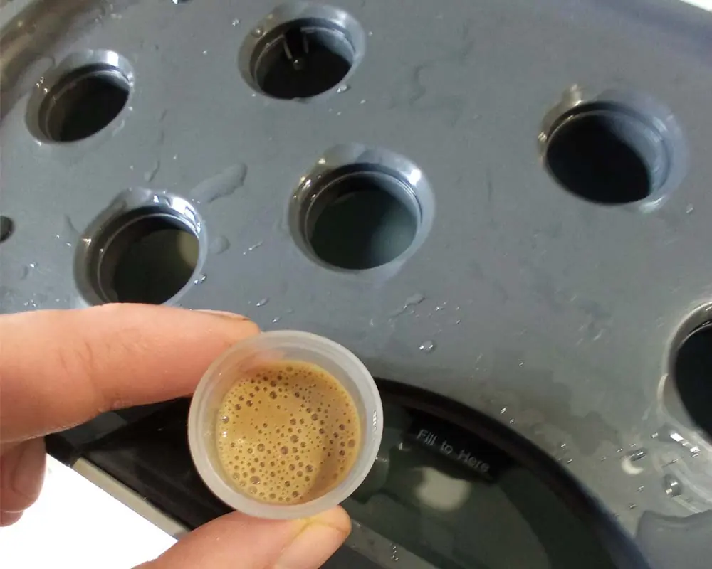 A small plastic measuring cup filled with a brown beneficial bacteria solution being added to an AeroGarden reservoir deck for root rot prevention.